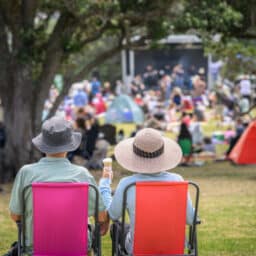 Couple sitting on camping chairs, unrecognizable crowds listen to the music concert in the park.