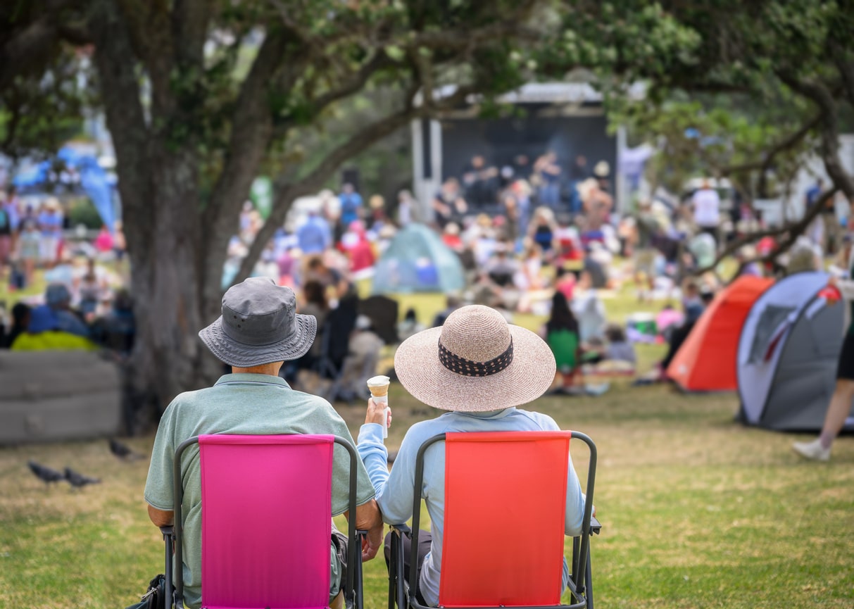 Couple sitting on camping chairs, unrecognizable crowds listen to the music concert in the park. 